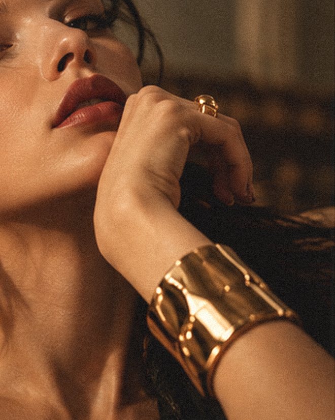 Mood image: a woman in low warm light, her hand at her jaw, wearing a broad gold cuff
